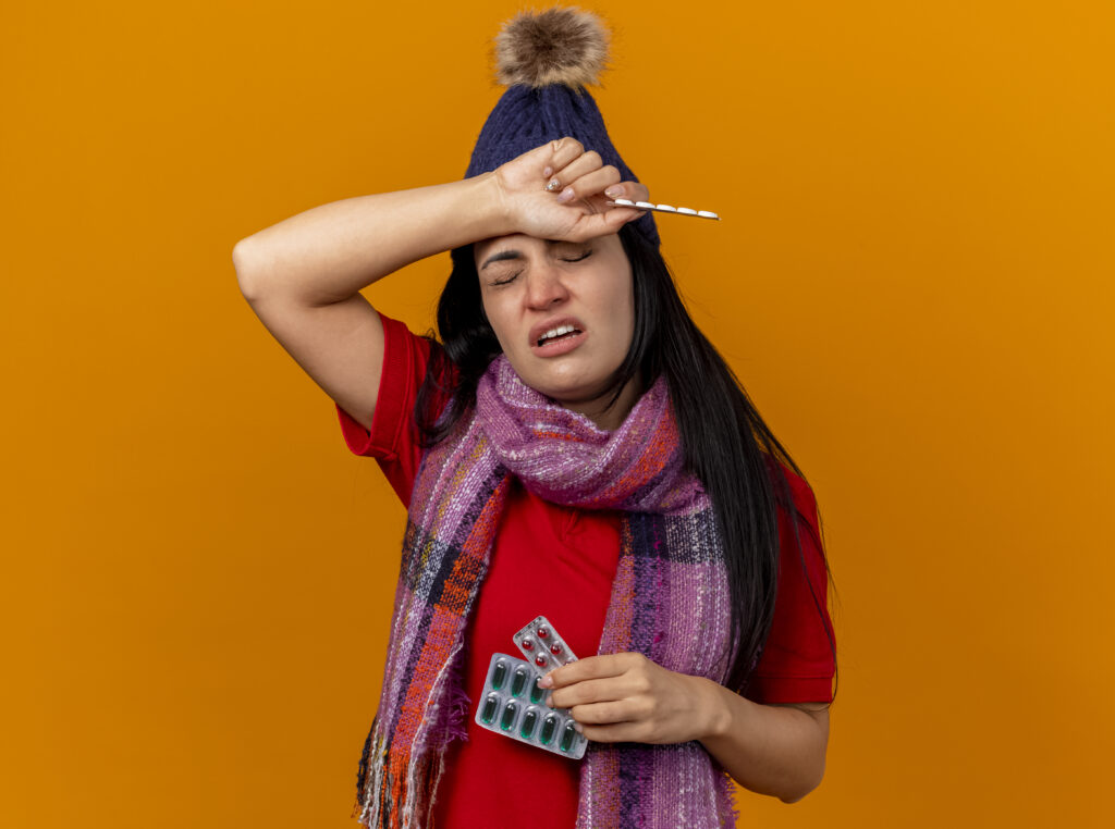 aching young caucasian ill girl wearing winter hat and scarf holding packs of capsules and tablets putting hand on head with closed eyes isolated on orange background with copy space