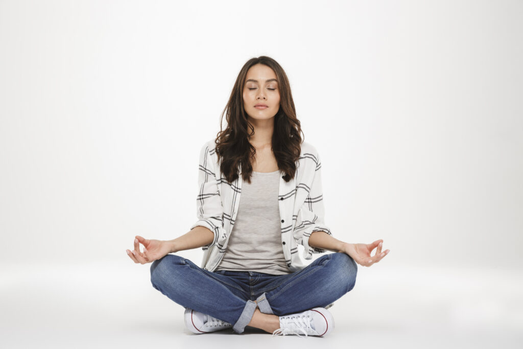 Full-length picture of concentrated woman in casual clothes meditating with closed eyes while sitting in lotus pose on the floor isolated over white wall - medstown