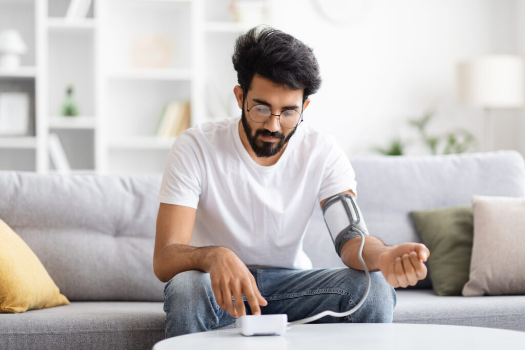 young Indian Guy Checking Blood Pressure With Upper Arm Monitor At Home, Millennial Eastern Man Sitting On Couch In Living Room And Measuring Arterial Tension With Medical Equipment, Copy Space