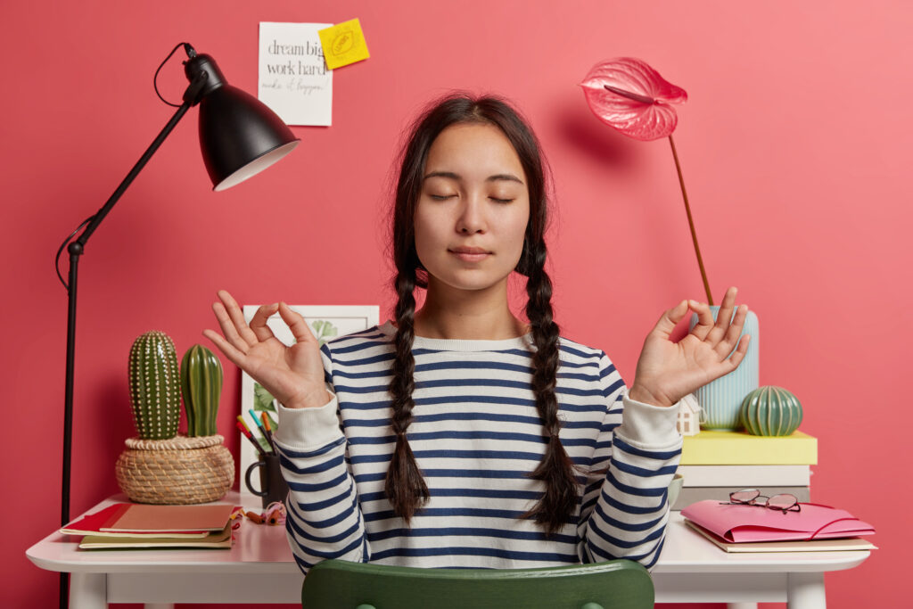 How to Make Your Heart 20 Years Younger Naturally 7 Relaxed Asian woman meditates at workplace, sits in zen pose against desktop with flowers, desk lamp, notepads, wears striped casual jumper, tries to relax after work, isolated over pink background