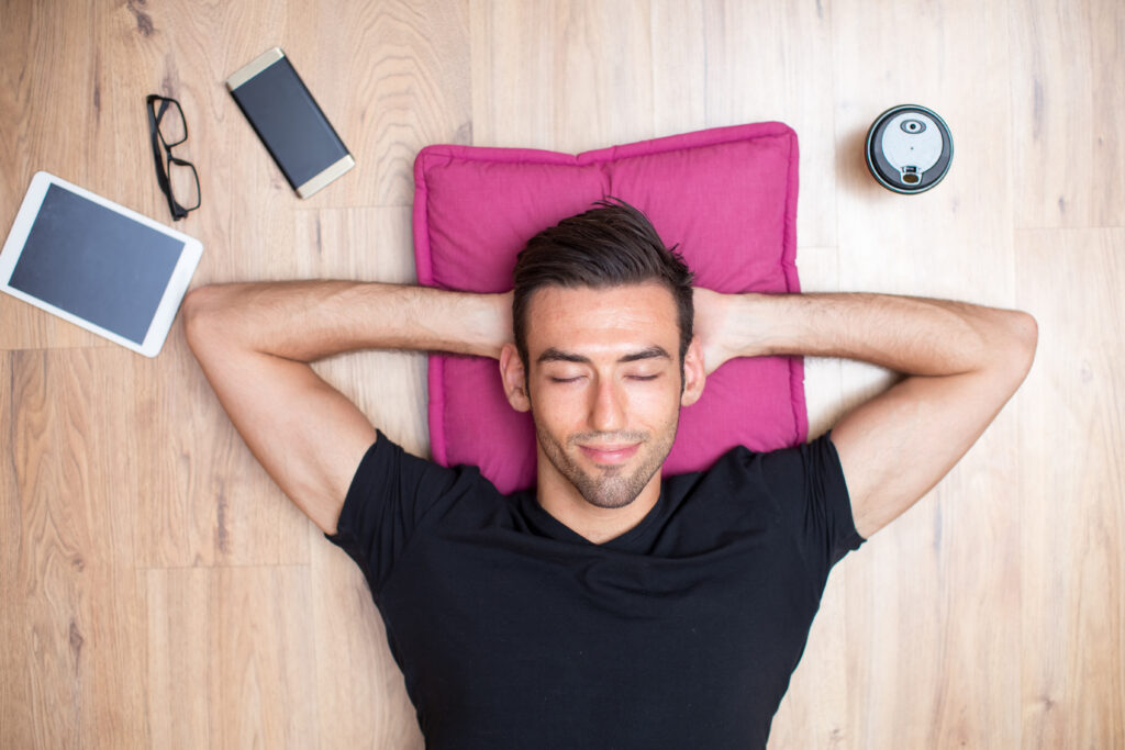 Closeup portrait of content young handsome man lying on floor with his hands behind head and eyes closed. Top view.