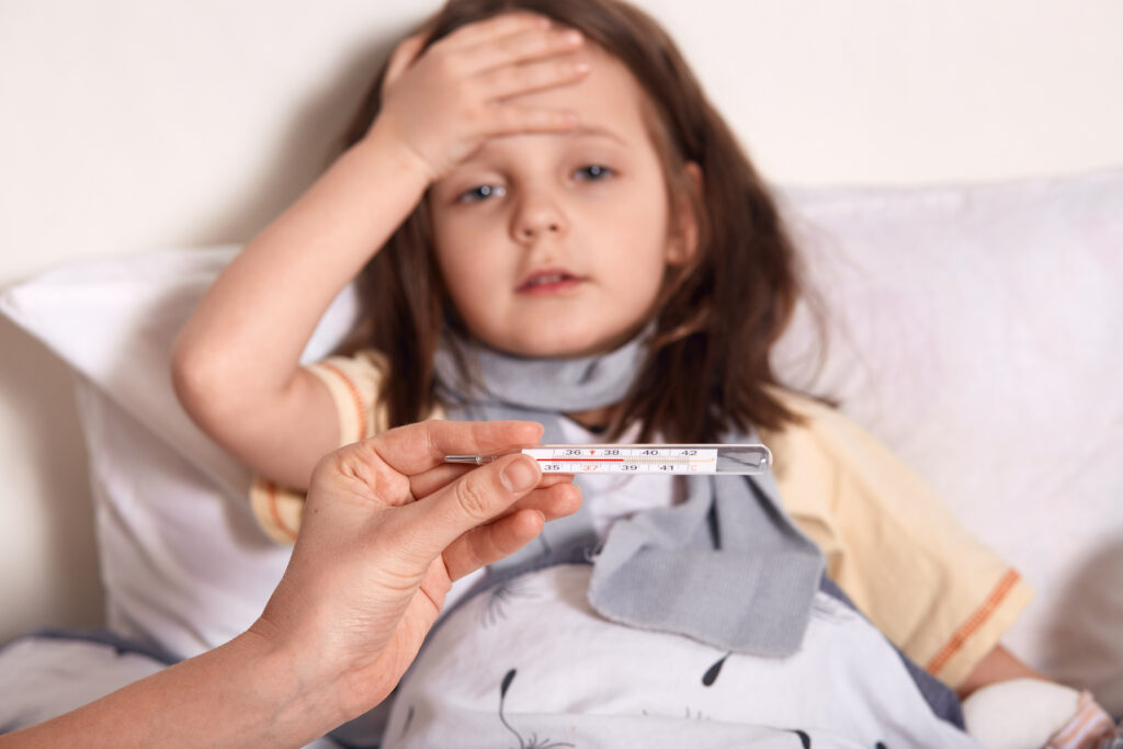 Mother holding thermometer in hands, sick little girl lying in bed and keeping her palm on forehead, looks sad and upset, suffering from high temperature, needs treatment and examining by doctor.