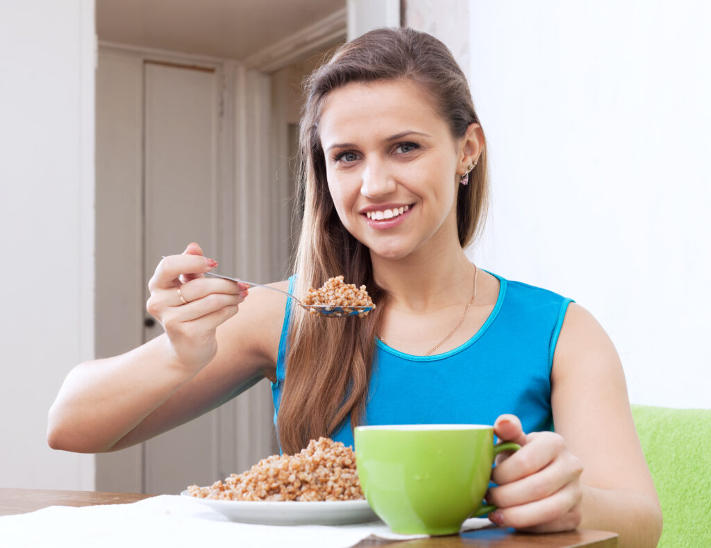 Smiling woman eats buckwheat cereal with spoon at home
